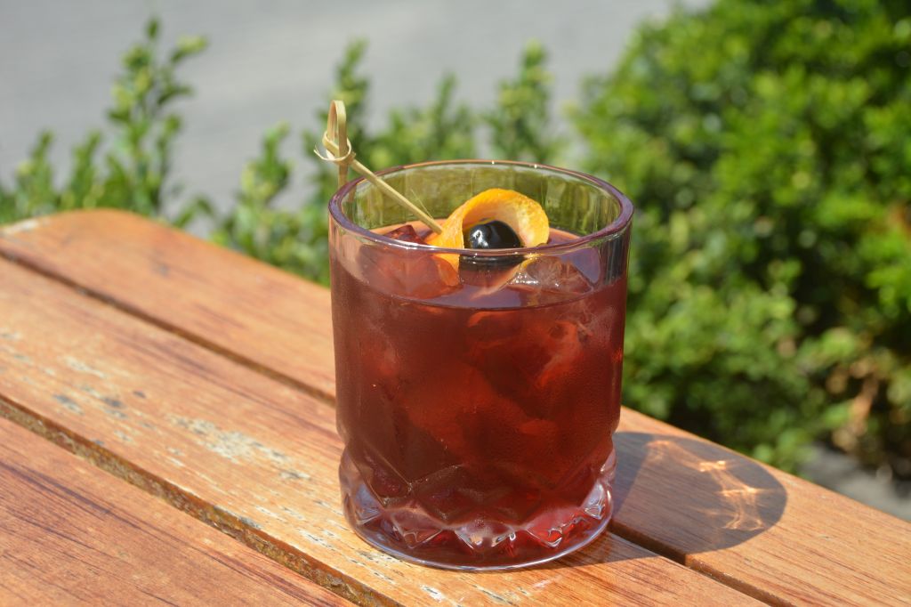 A glass of red cocktail garnished with an orange slice and a black olive, set on a wooden table against a backdrop of lush greenery.