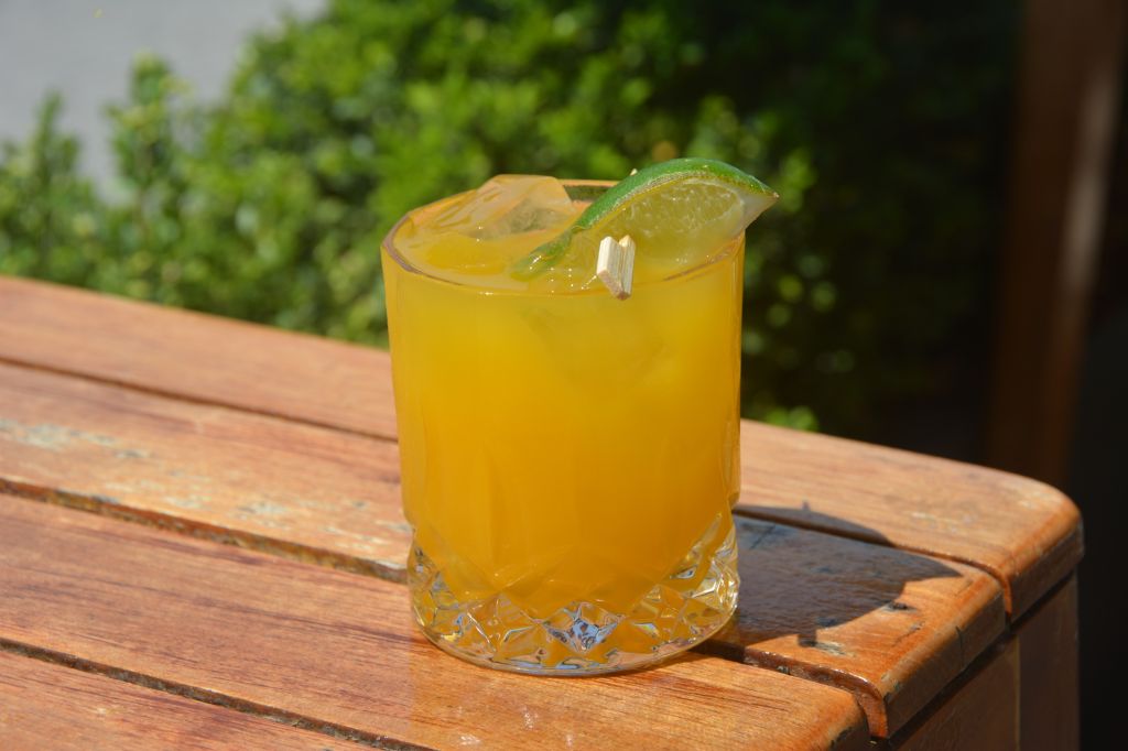 A glass of orange-colored beverage garnished with a lime slice on a wooden table against a blurred green background.