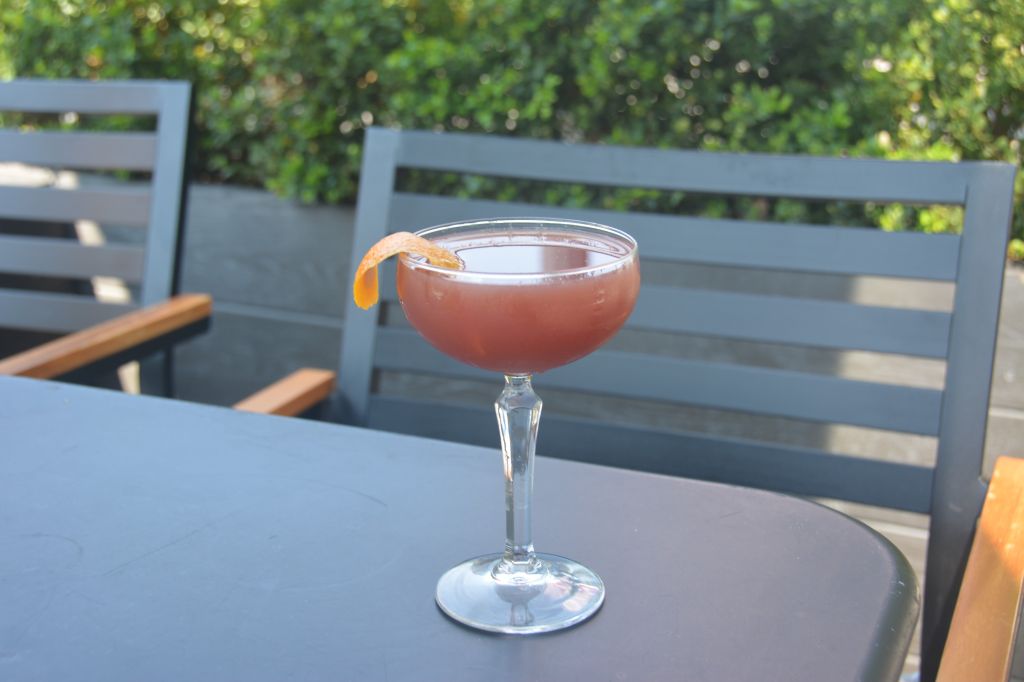 A glass of pink cocktail garnished with an orange slice, sitting on a table with a wooden bench in a lush, green outdoor setting.