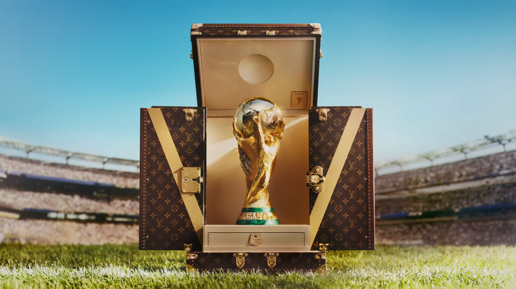 A golden trophy representing the FIFA World Cup sits inside a Louis Vuitton-branded travel case on a grassy field, with a stadium visible in the background.