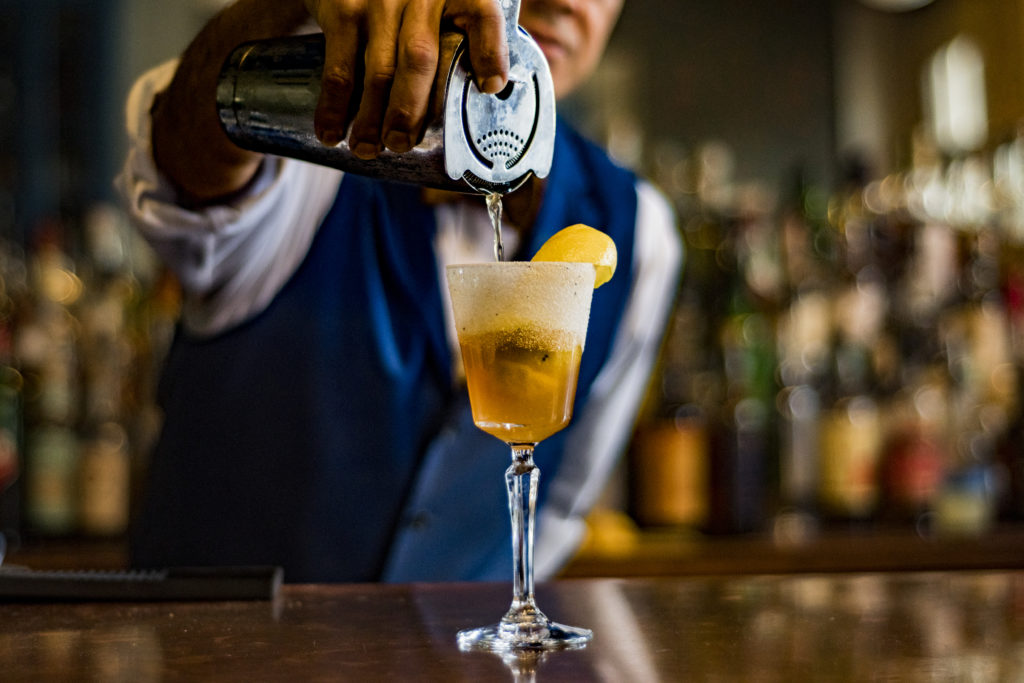 A bartender pouring a glass of amber-colored beverage, likely a cocktail, with a lemon slice garnish.