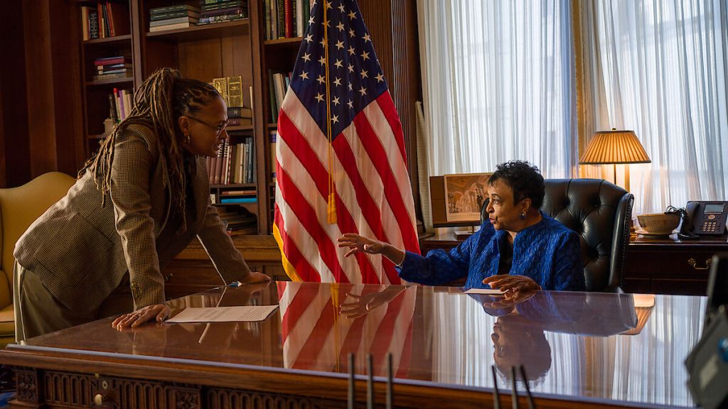 Two people, one in a blue jacket and one with dreadlocks, discussing in an office with an American flag.