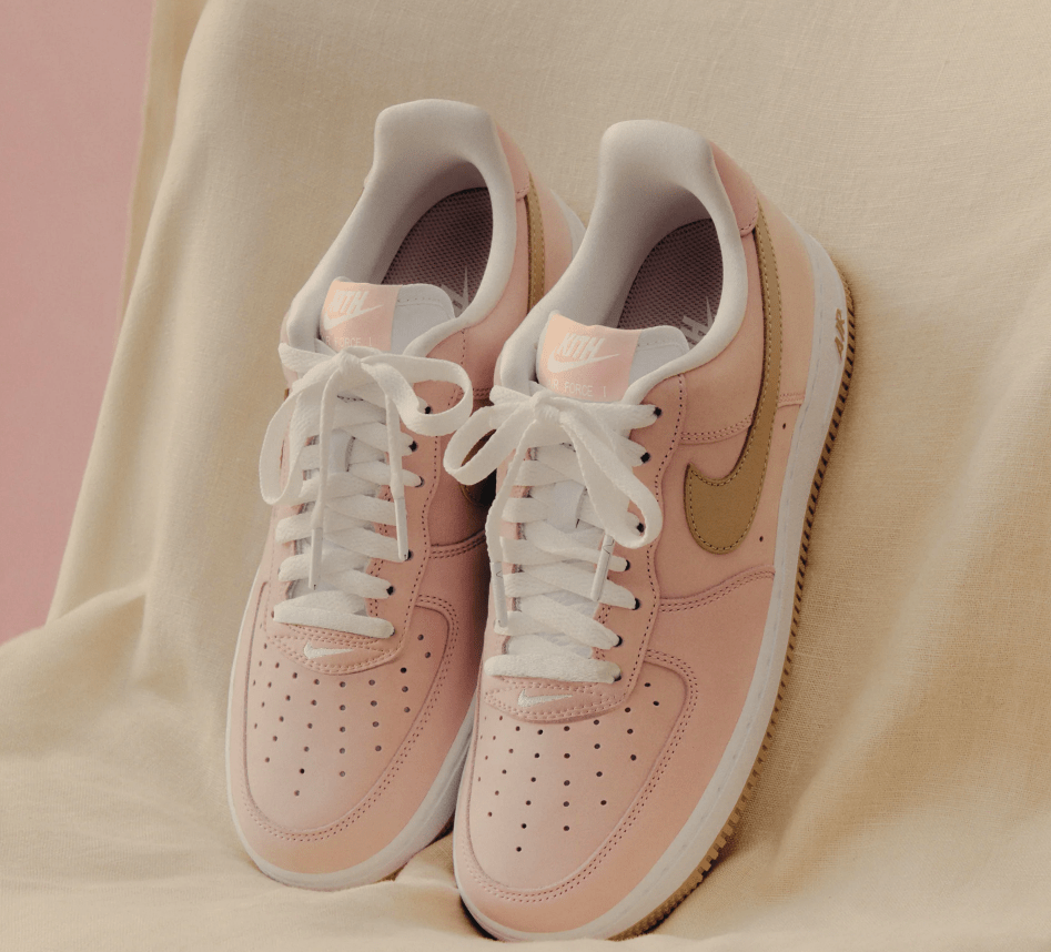 A pair of light pink Nike Air Force 1 sneakers with white soles and laces, resting on a light-colored fabric.