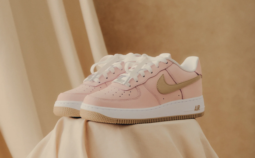 A pair of pink and white Nike Air Force 1 sneakers with a gum sole, resting on a beige fabric surface.