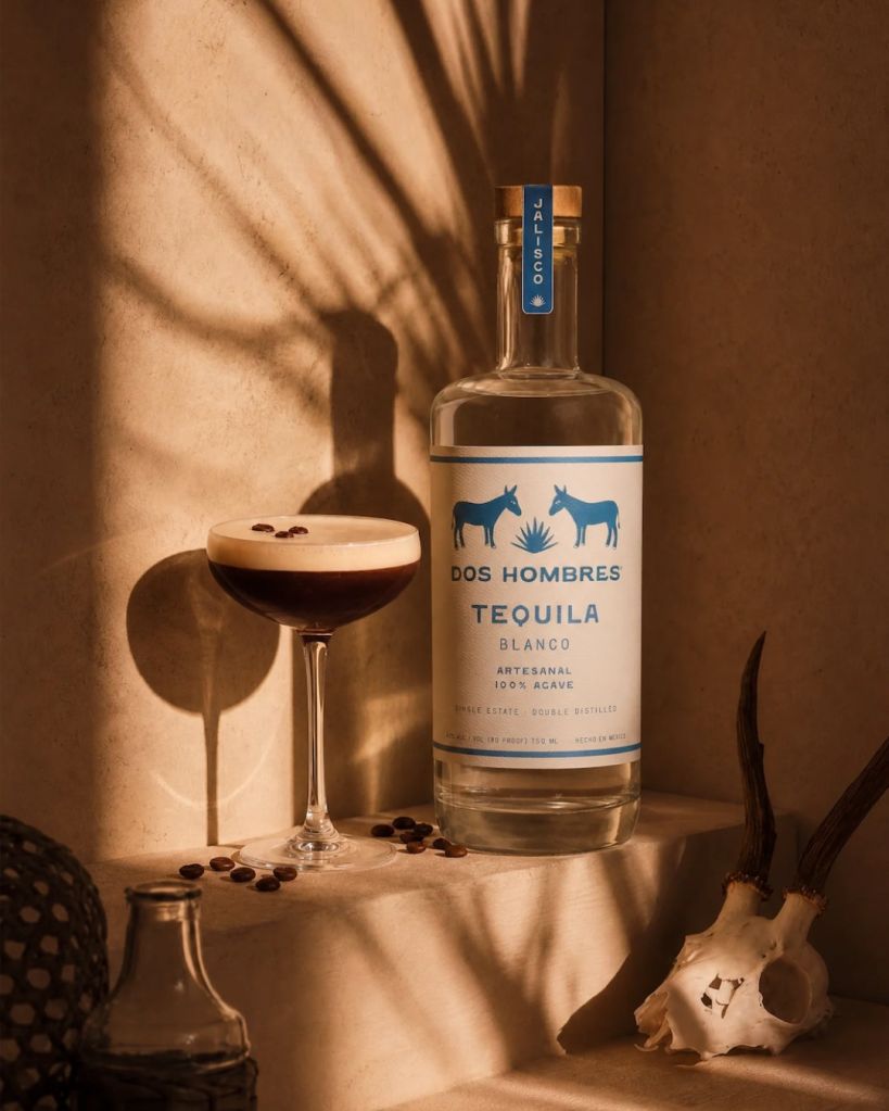 A bottle of Dos Hombres Tequila Blanco on a table with a glass of tequila cocktail and coffee beans, with dramatic lighting casting shadows on the wall.