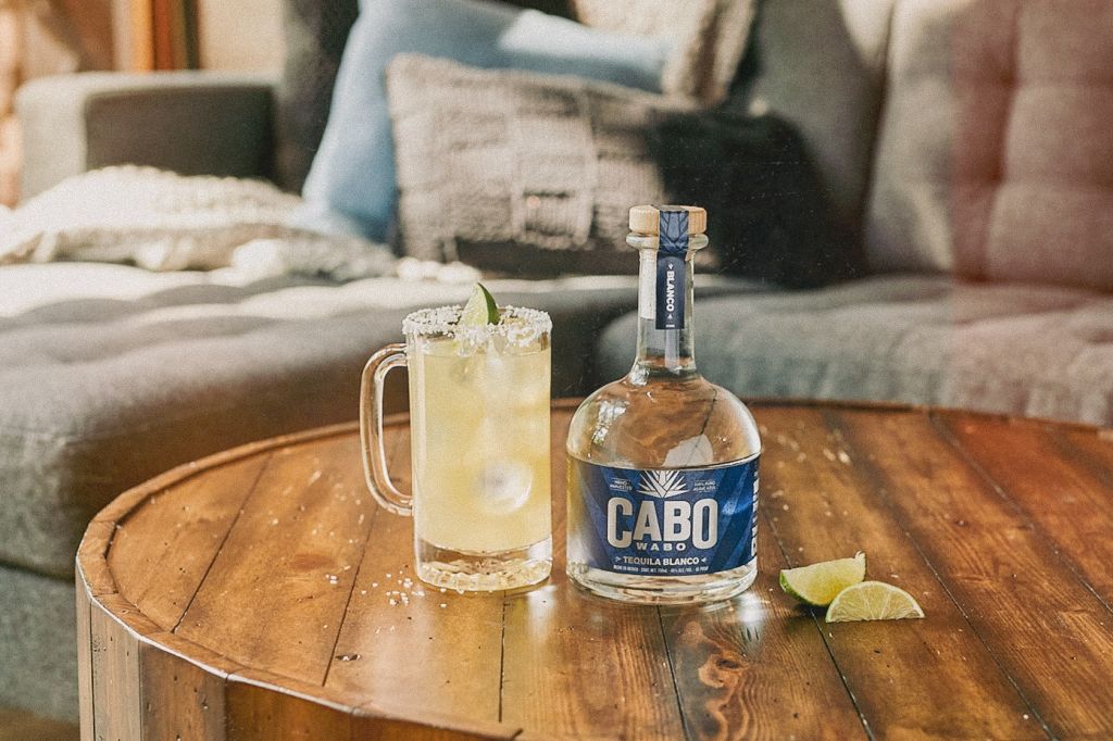 A wooden table with a glass of lemonade and a bottle of Cabo tequila blanco.
