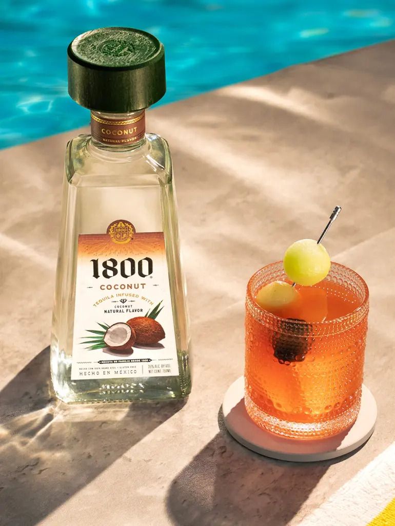 A bottle of 1800 Coconut tequila and an orange cocktail with a lemon garnish, set against a turquoise pool.