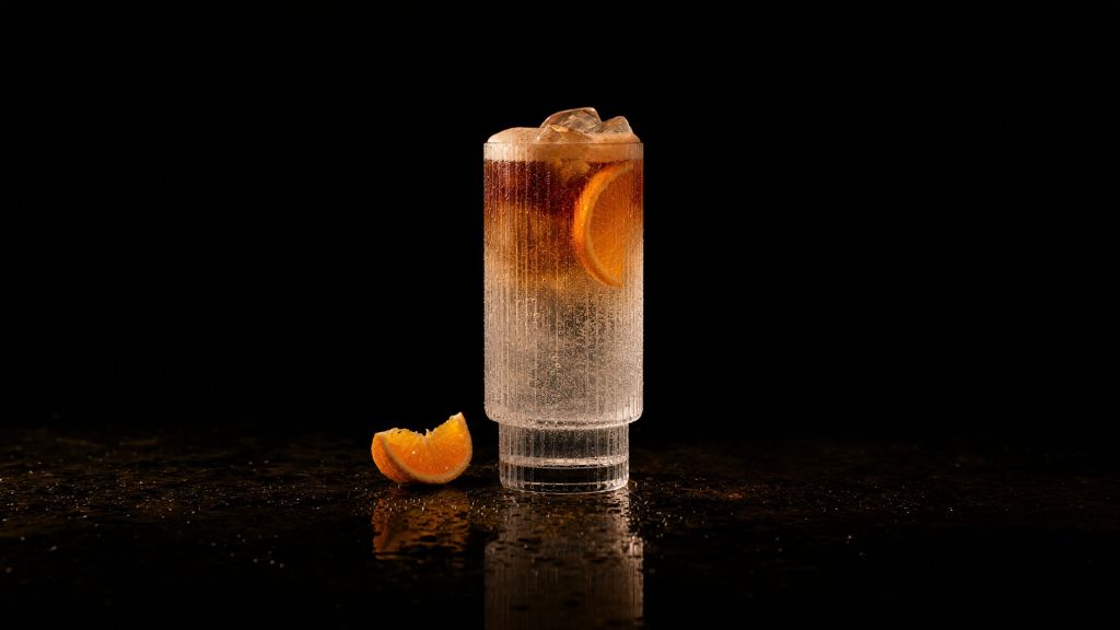A glass of orange cocktail with ice cubes and a slice of orange garnish on a dark reflective surface.