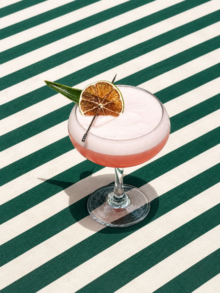 A glass cocktail with a slice of dried orange garnish on a green and white striped background.