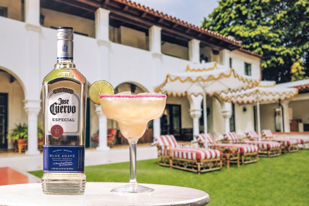A bottle of Jose Cuervo Especial tequila and a margarita cocktail on a table in front of a Spanish-style building with arched windows and a tiled roof, surrounded by outdoor seating.
