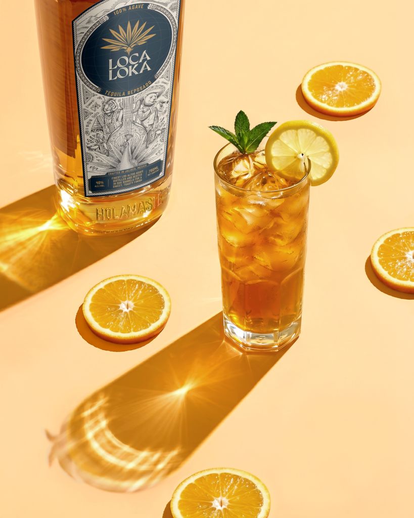 A glass of orange cocktail with lemon and mint garnish, next to a bottle of Loca Loka tequila on a yellow background.