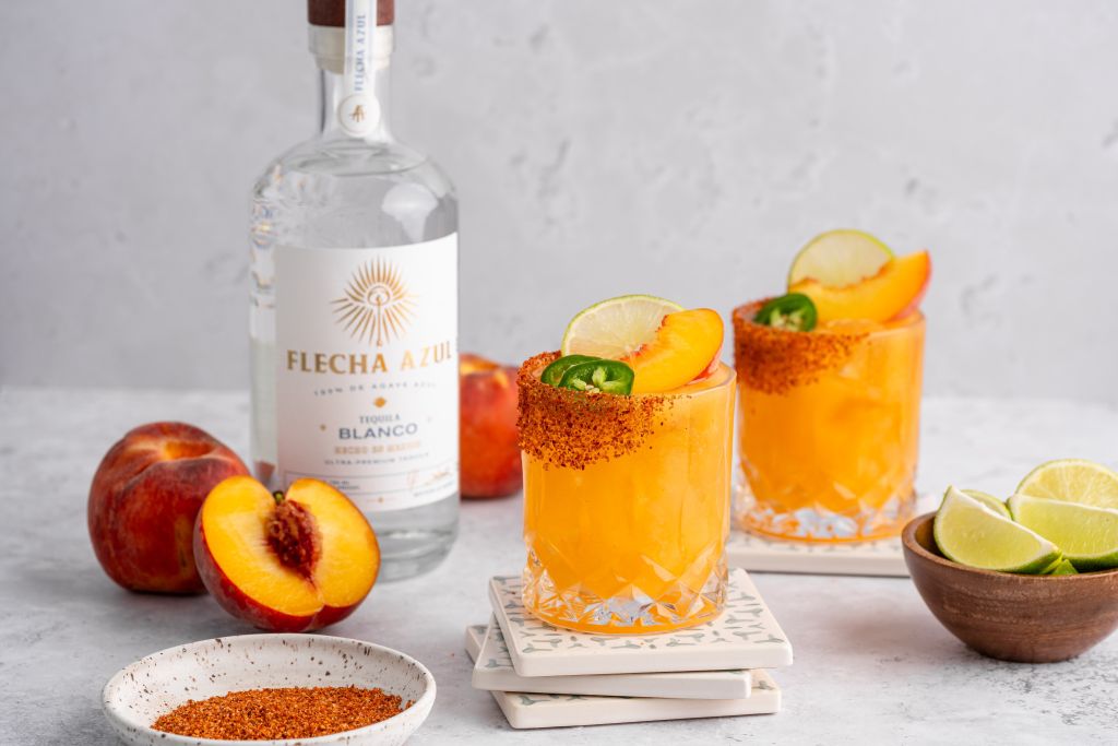 A bottle of Flecha Azul Blanco tequila, orange cocktails garnished with lime and jalapeño, and fresh peach slices on a marble surface.