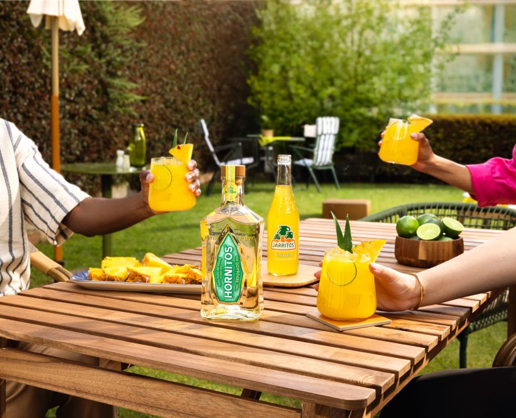 Outdoor patio with wooden table, glasses of orange drink, bottles of Hornitos tequila and Jarritos soda, and hands holding the drinks.