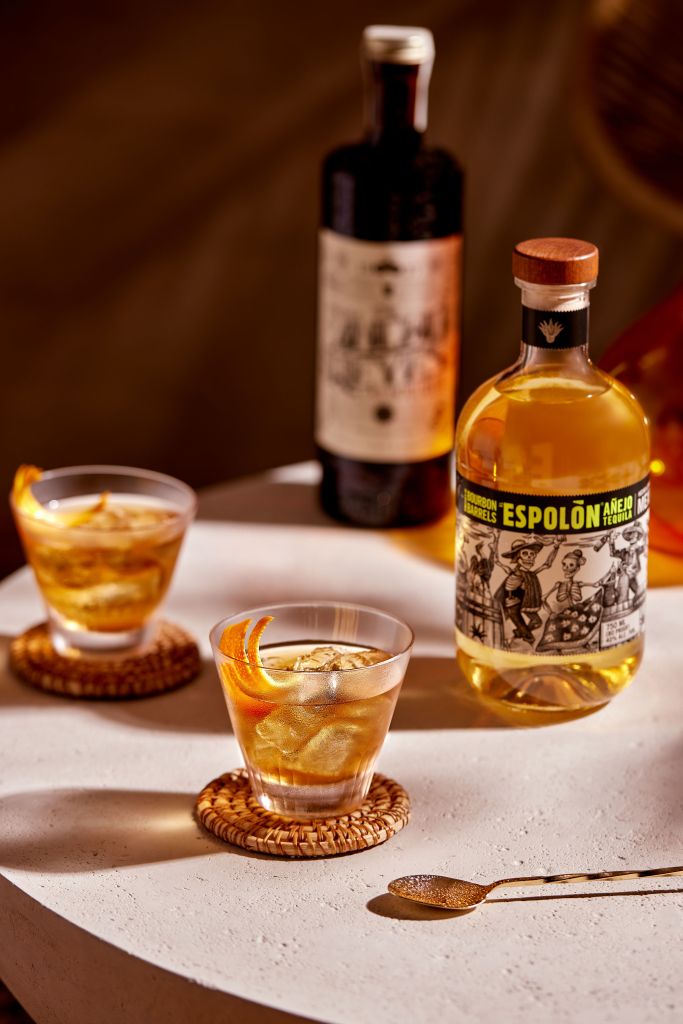 A bottle of Espolon tequila and a glass of amber-colored cocktail on a woven coaster, with a bottle of red wine in the background.