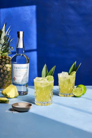 Two glasses of pineapple cocktail with Teremana tequila bottle and fresh pineapple slices on a blue background.