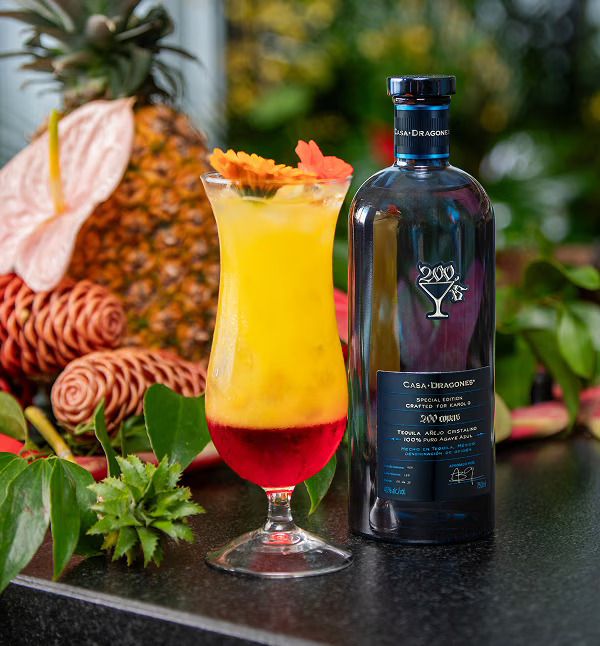 A tropical cocktail with pineapple and orange slices, served in a glass next to a bottle of Casa Dragones tequila against a lush, green background.