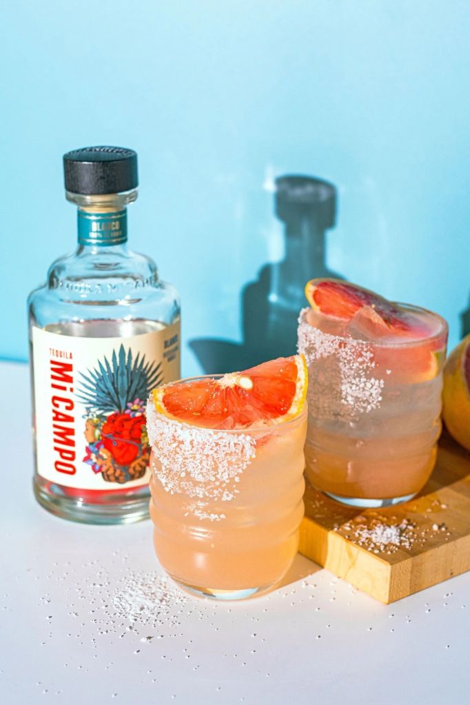 Two glasses of orange cocktail garnished with orange slices, alongside a bottle of Mi Campo tequila on a light blue background.