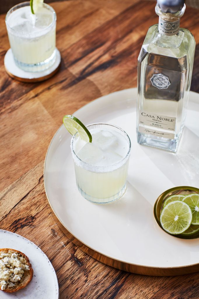 A glass of margarita with lime garnish on a wooden table, next to a bottle of Casa Noble tequila.