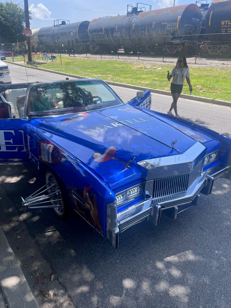 A blue police car with "MARLII" written on the side is parked on the street, with a person walking in the background and train cars on the tracks behind them.