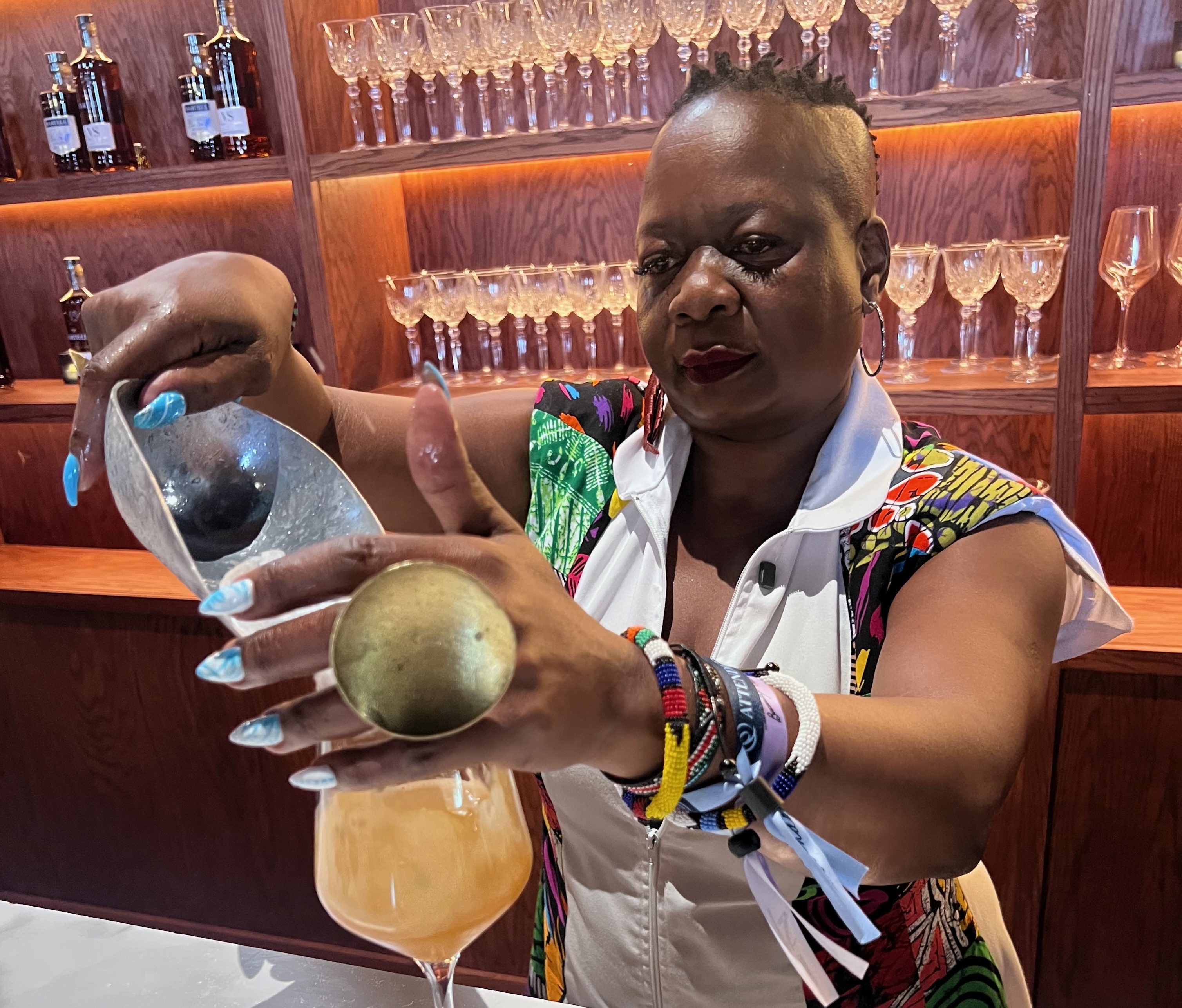 A person wearing colorful clothing and jewelry is holding a glass of orange liquid and a lime wedge at a bar with shelves of glassware behind them.