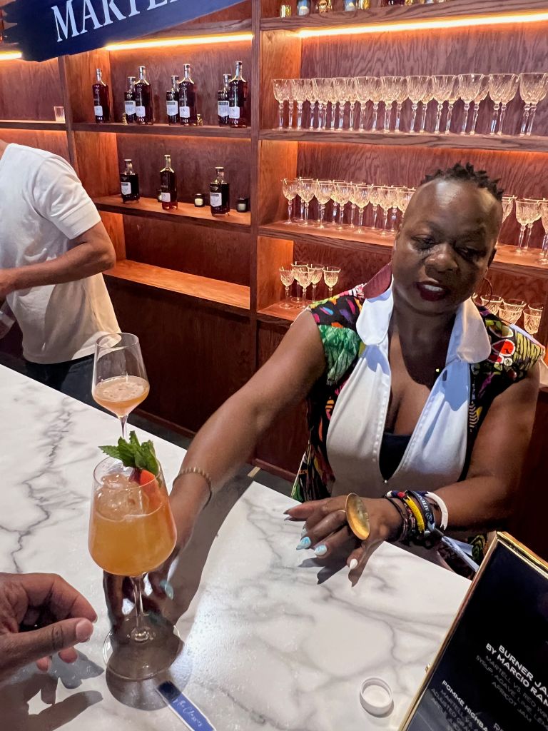 A person in a colorful outfit sitting at a bar, with shelves of wine glasses and bottles behind them.