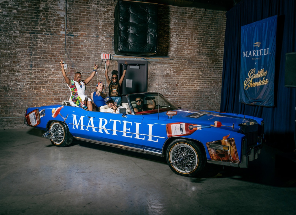 A group of people riding in a blue Martell convertible car, with Martell branding visible on the vehicle.