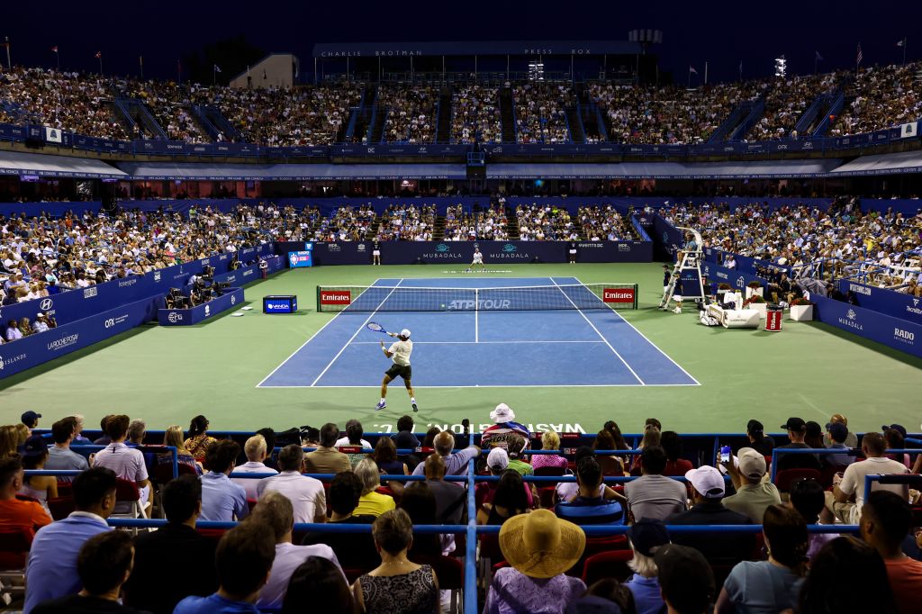 A crowded tennis stadium at night, with a player in white clothing hitting a tennis ball on the court. The stadium is filled with spectators in the stands.