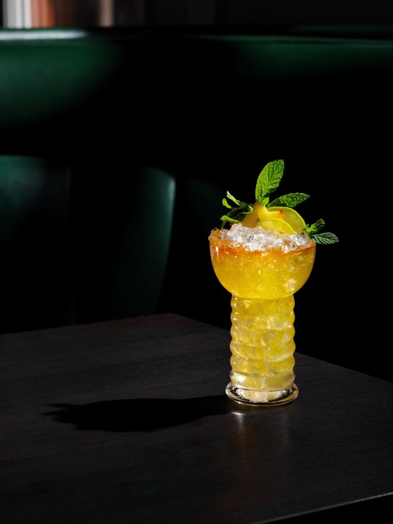 A glass filled with a yellow, frothy cocktail garnished with a lemon slice and mint leaves, set against a dark background.