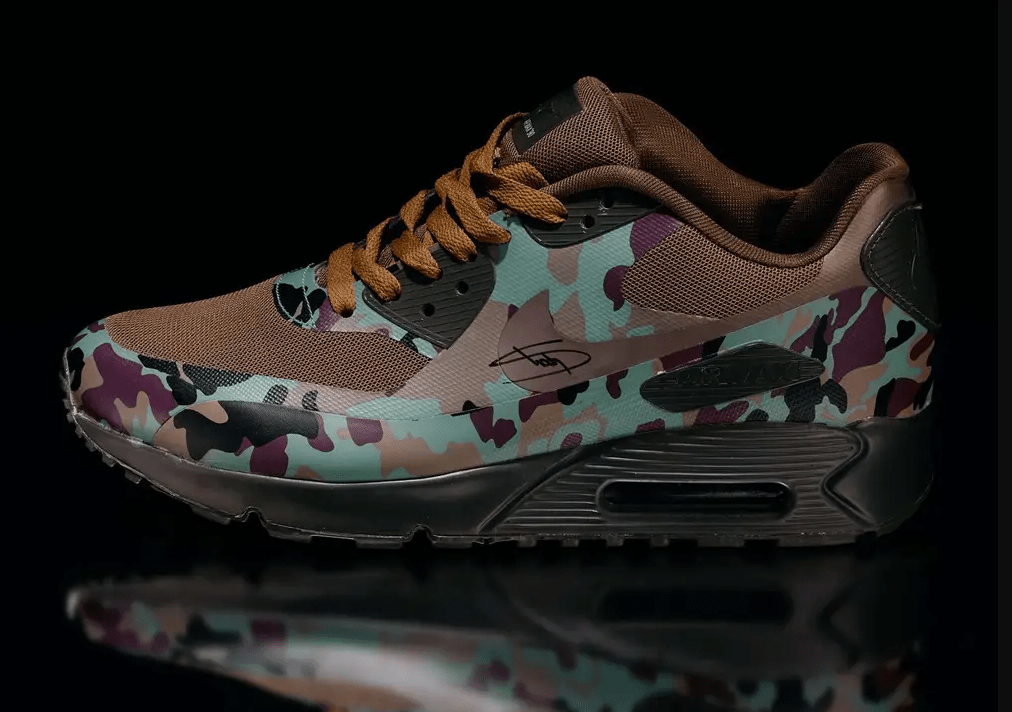 Camouflage-patterned Nike Air Max 90 sneaker with brown mesh and laces against a dark background.