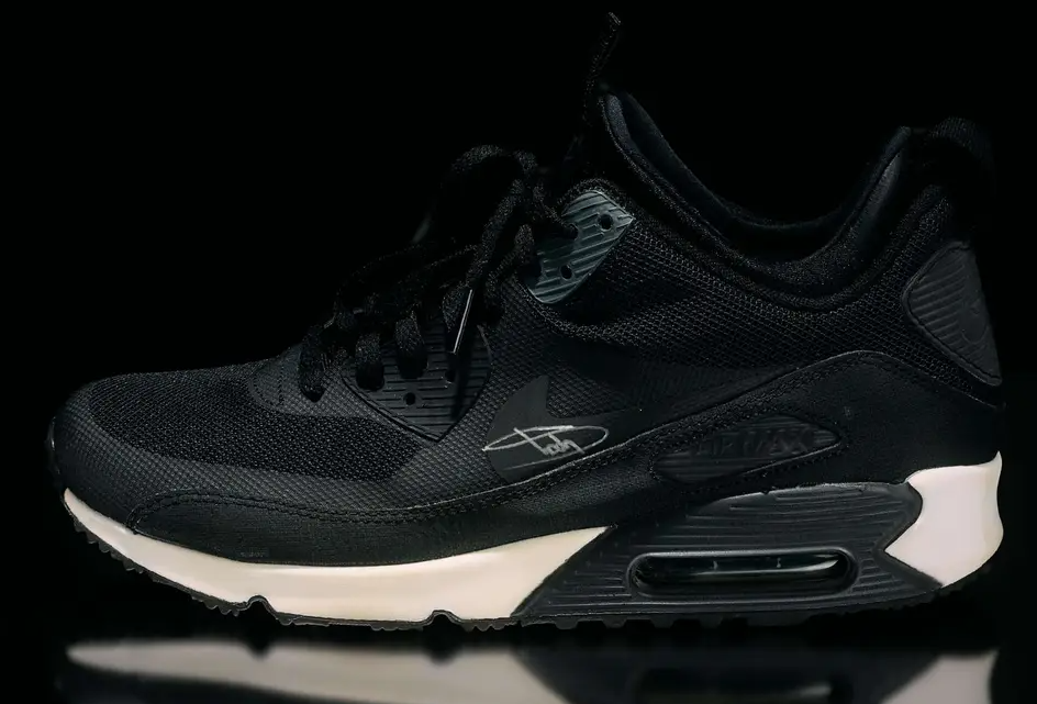 Black Nike Air Max 90 sneakers with white soles against a dark background.