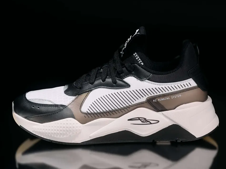 A black and white athletic shoe with the text "RS Running System" and a white midsole on a reflective surface.