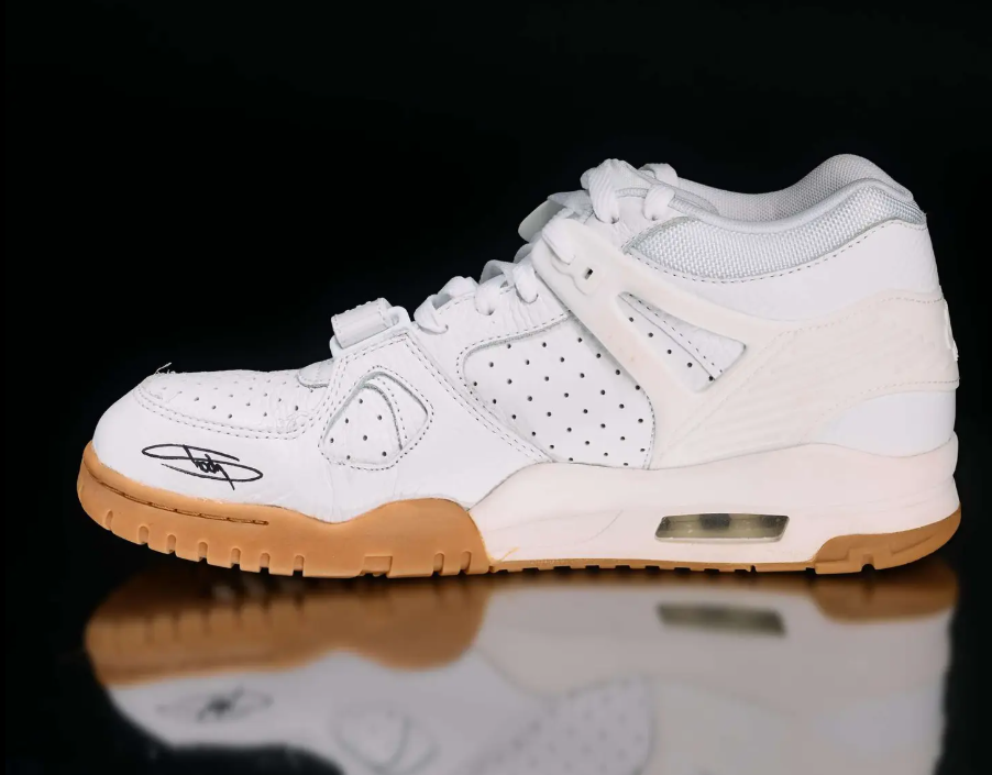 A white athletic shoe with perforated details and a gum sole, reflected on a shiny surface against a dark background.