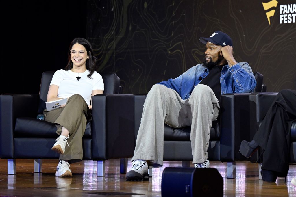 Two people, a woman and a man, sitting on stage at a Fanatics Fest event. The woman is smiling and the man is wearing a denim jacket.