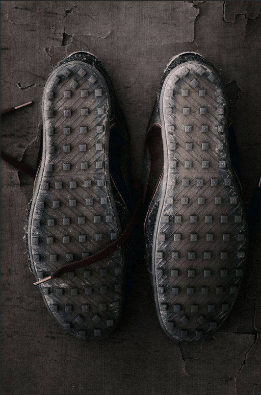 A pair of worn, textured sneakers with a checkered pattern on the soles, resting on a dark, cracked surface.