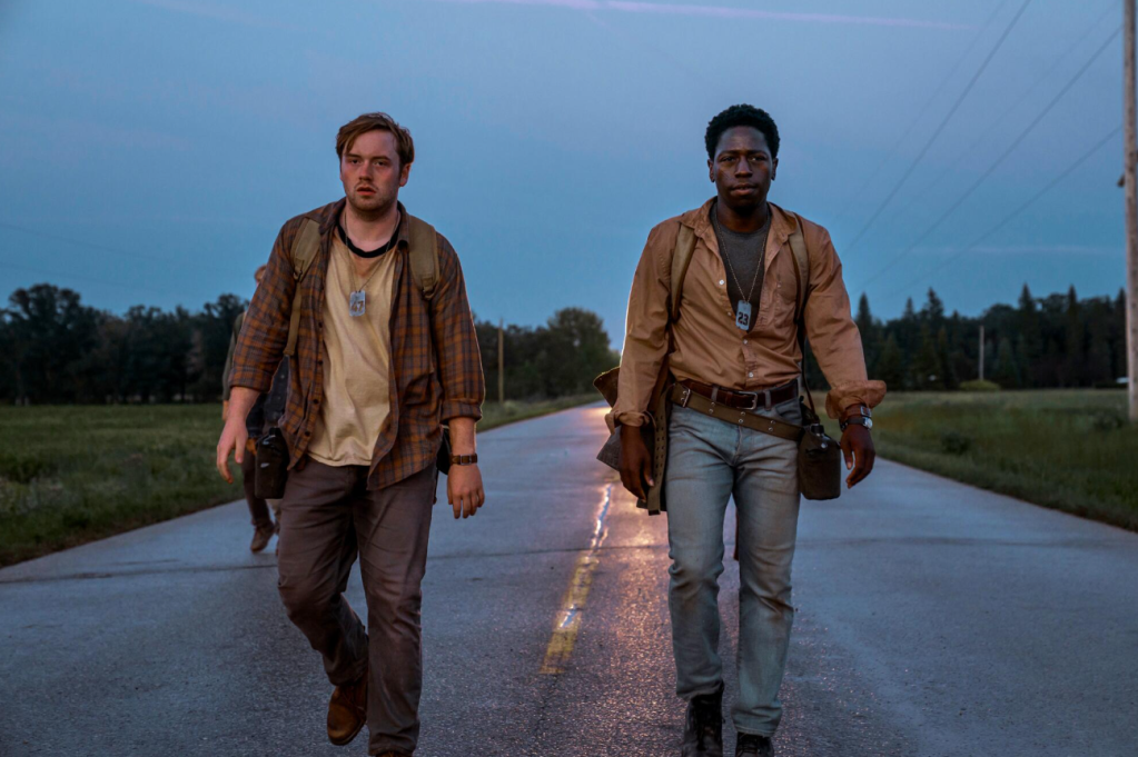 Two men walking down a rural road at dusk, one wearing a plaid jacket and the other a tan jacket.