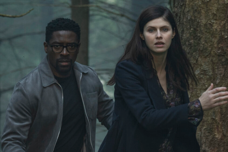 Two people, a man and a woman, standing in a wooded area. The woman is wearing a dark coat and the man is wearing glasses.