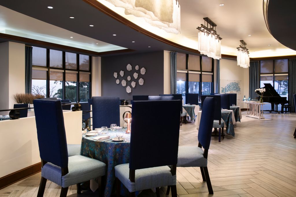 A modern, upscale dining room with dark walls, large windows, and a grand chandelier. Several tables are set with place settings and a grand piano is visible in the background.