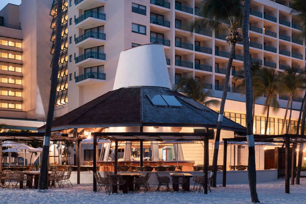 Beachfront resort with palm trees, illuminated outdoor dining area, and multi-story hotel building in the background.