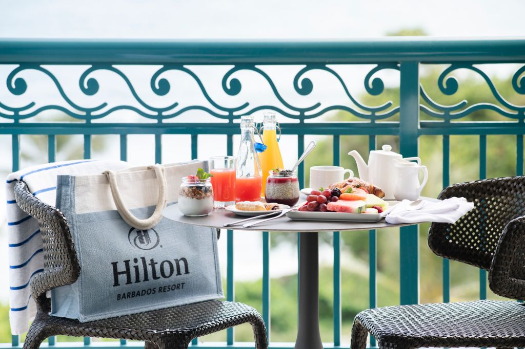 A table set with a breakfast spread, including a Hilton Barbados Resort tote bag, on a balcony overlooking a scenic view.