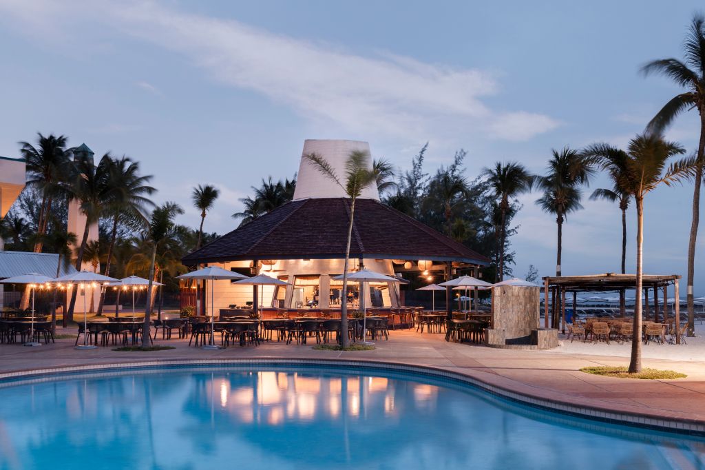 Tropical resort with palm trees, pool, and open-air restaurant at dusk, with lights reflecting in the water.