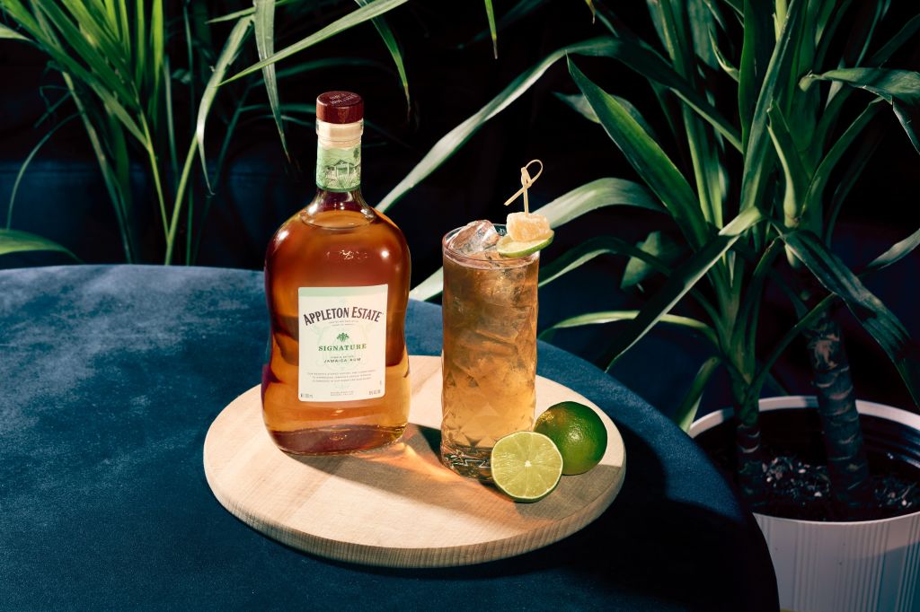 A bottle of Appleton Estate Signature rum and a glass of mixed drink with lime garnish, set against a backdrop of lush green foliage.