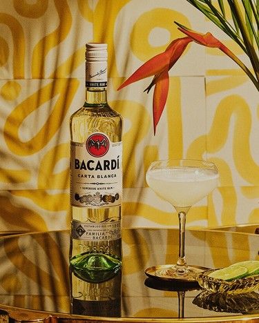 A bottle of Bacardi rum and a cocktail glass on a table with a tropical flower and yellow and orange abstract background.