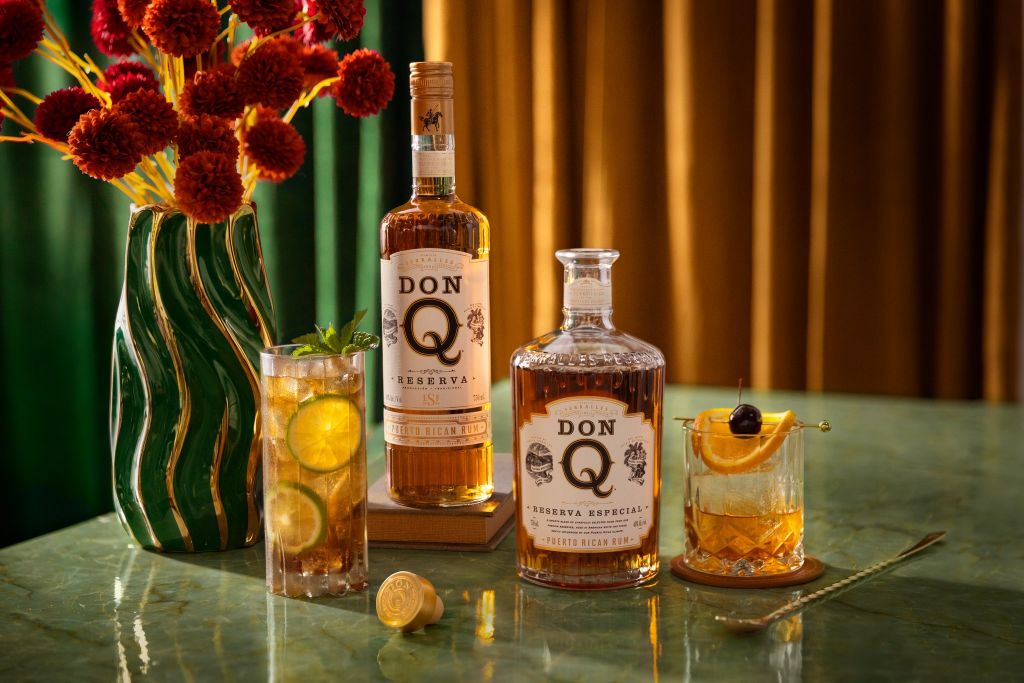 Two bottles of Don Q rum, a glass of cocktail, and a vase of red flowers on a wooden table.