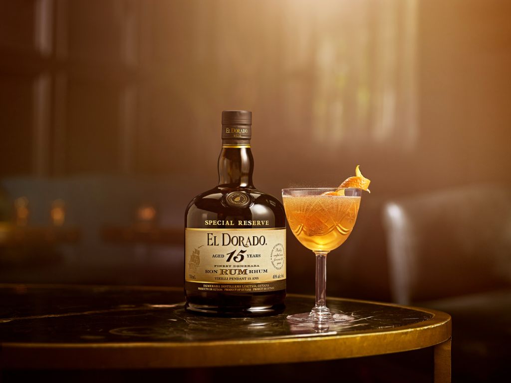 A bottle of El Dorado 15-year-old rum and a glass of an orange cocktail on a wooden barrel in a dimly lit setting.