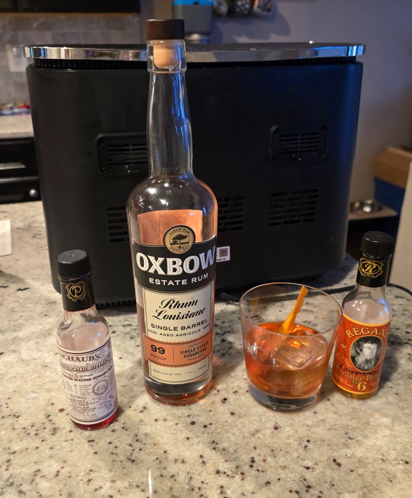 A bottle of Oxbow Estate Rum, a glass of Regan's Orange Bitters, and a glass of Campari on a granite countertop with a black refrigerator in the background.