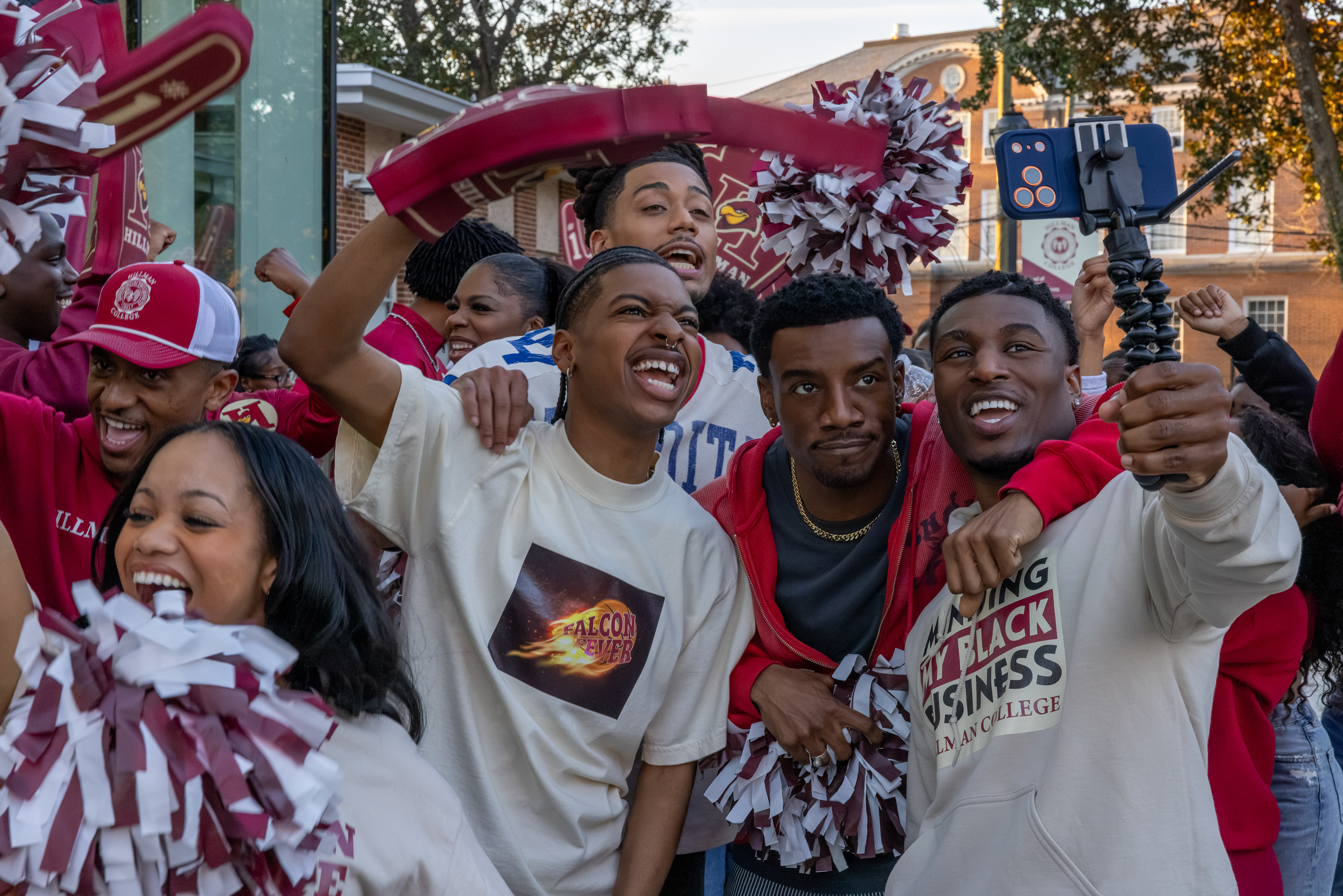 "College students at a lively outdoor event, smiling and cheering with pom-poms and team gear, taking selfies and enjoying a festive, energetic atmosphere on campus."