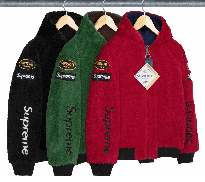 Three Supreme-branded hooded jackets in black, green, and red, hanging on wooden hangers against a gray background.