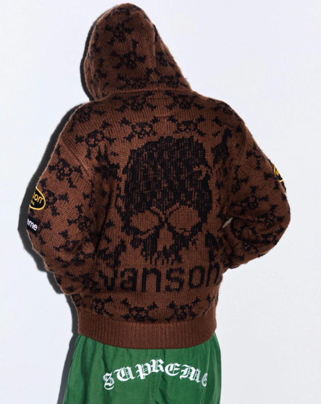A person wearing a hooded brown sweater with a skull graphic and the word "Supreme" printed on it.
