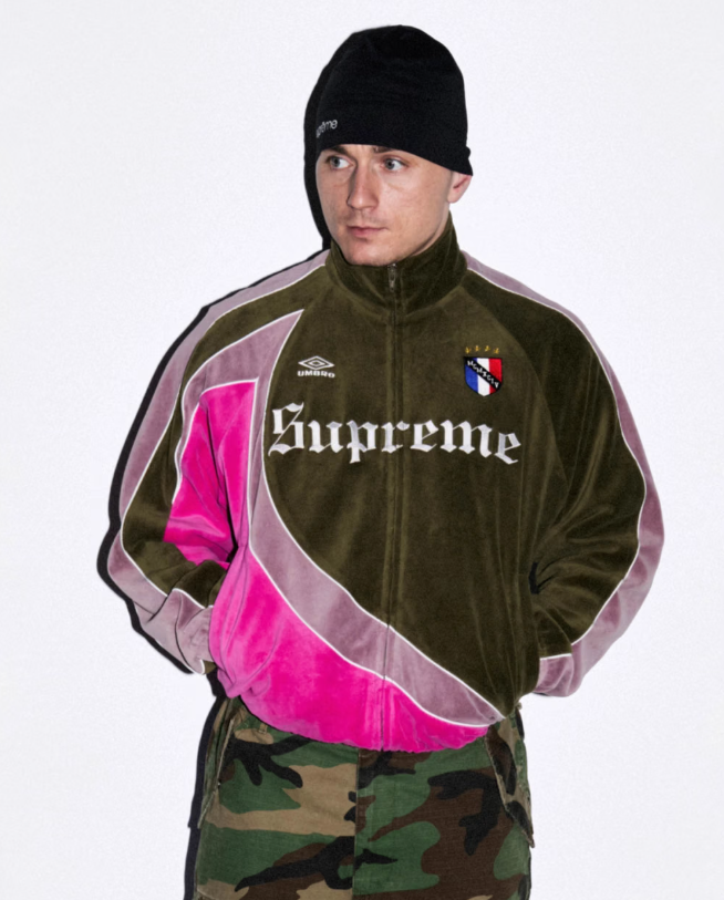 A person wearing a black beanie and a green and pink Supreme-branded jacket with camouflage pants.
