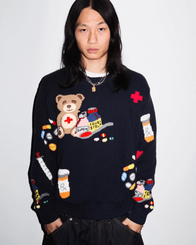 A person wearing a black sweater with a teddy bear and medical supplies printed on it, standing against a plain white background.